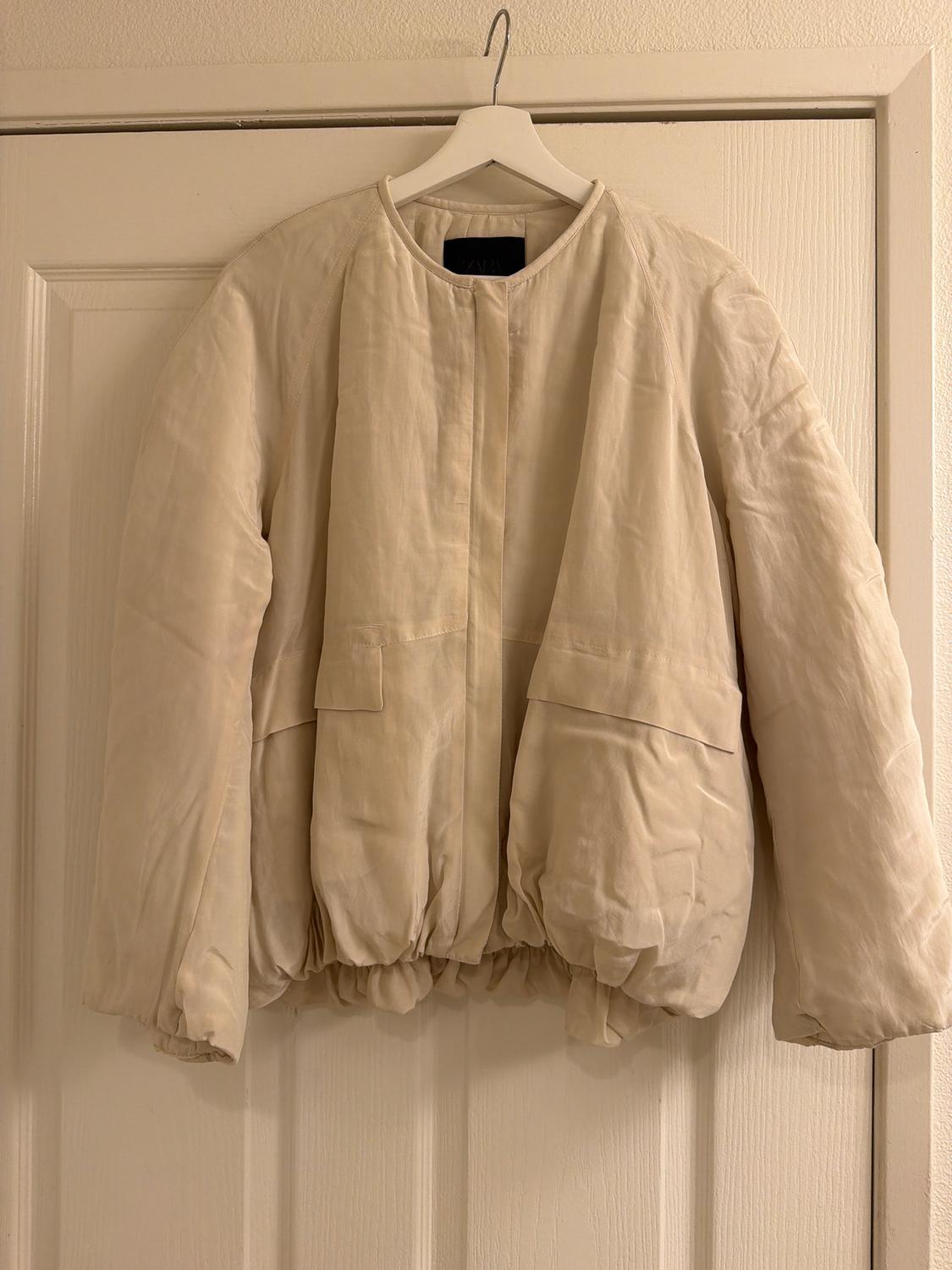 Beige quilted jacket hanging on a white hanger over a door. Simple design with front pockets.