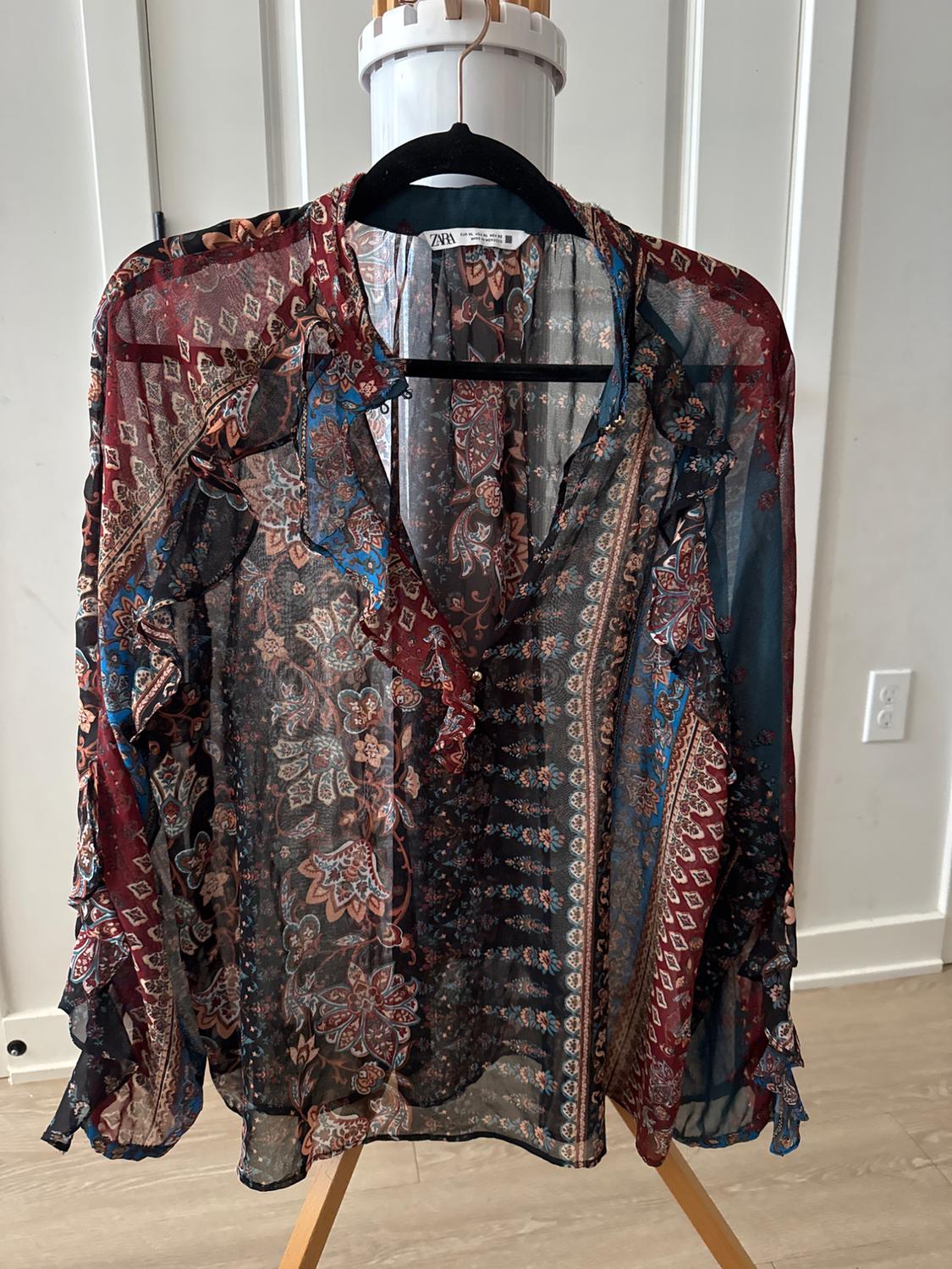 A sheer blouse with a floral and geometric print in shades of red, blue and brown, hanging on a black hanger.
