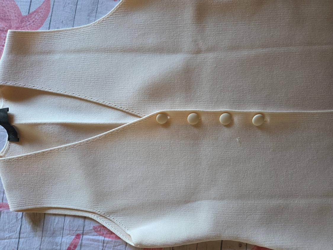 Beige knitted V-neck waistcoat with four buttons, laid out on a wooden surface with pink details. Relaxed style.