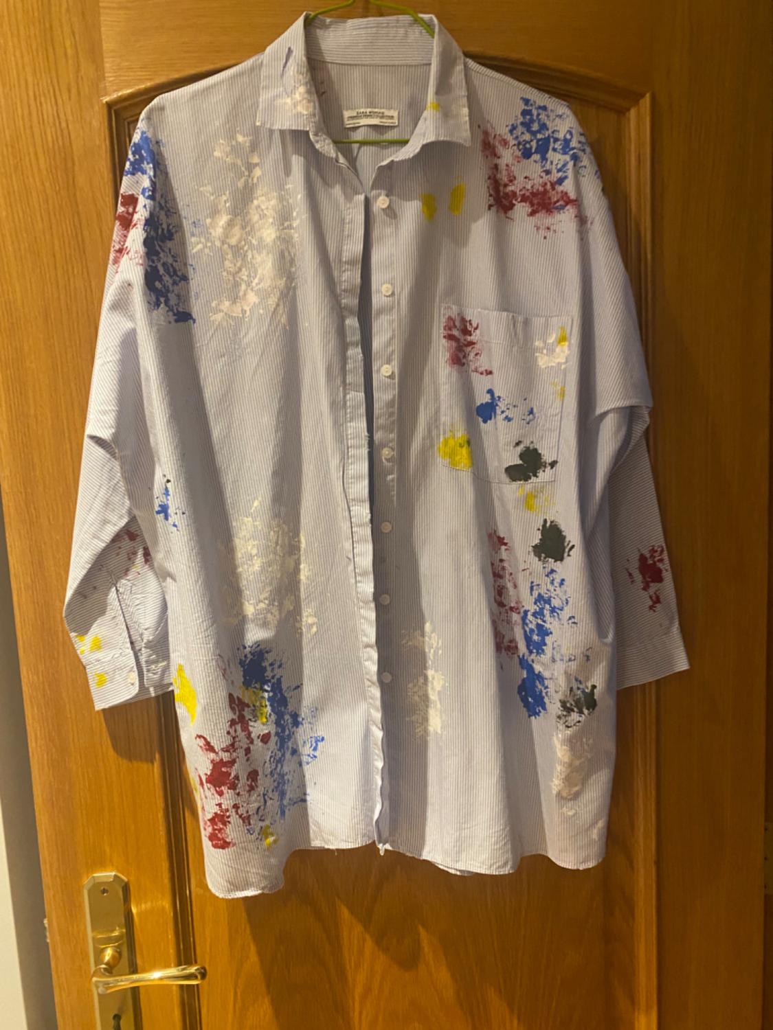 Front view of a blue striped shirt with multicoloured paint splatters and a front pocket.