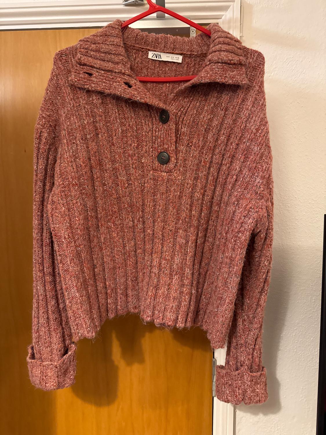 Chunky terracotta knit sweater with a high neck and two large buttons, hanging on a red hanger in front of a wooden door.