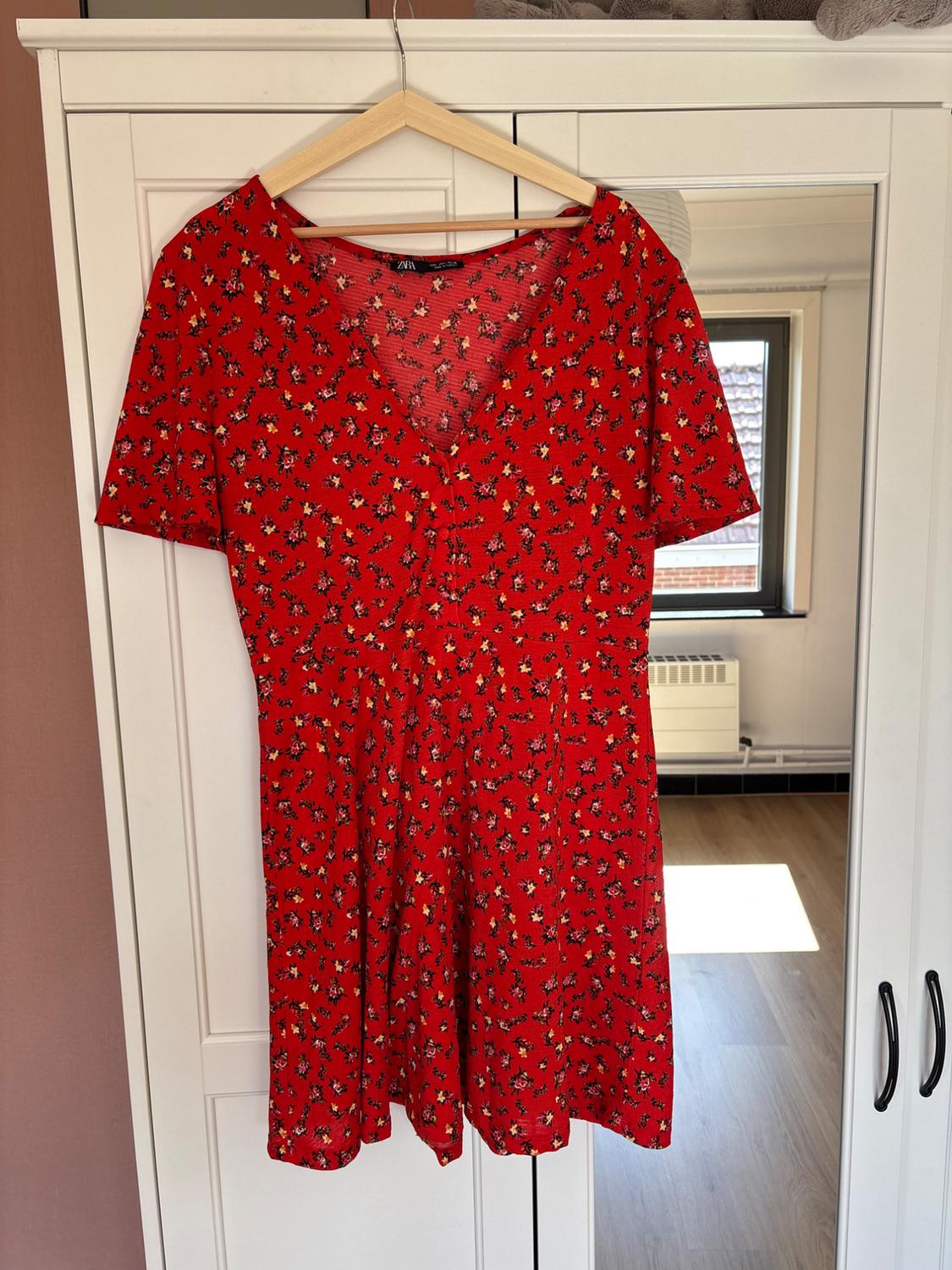 Red short dress with a V-neck and floral print, hanging on a hanger in front of a white wardrobe with a mirror. Text: ZARA.