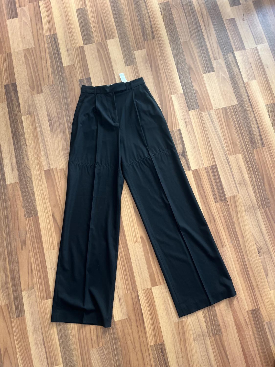 Black wide-leg trousers on a light wooden floor with a geometric pattern. Visible label on the waistband.