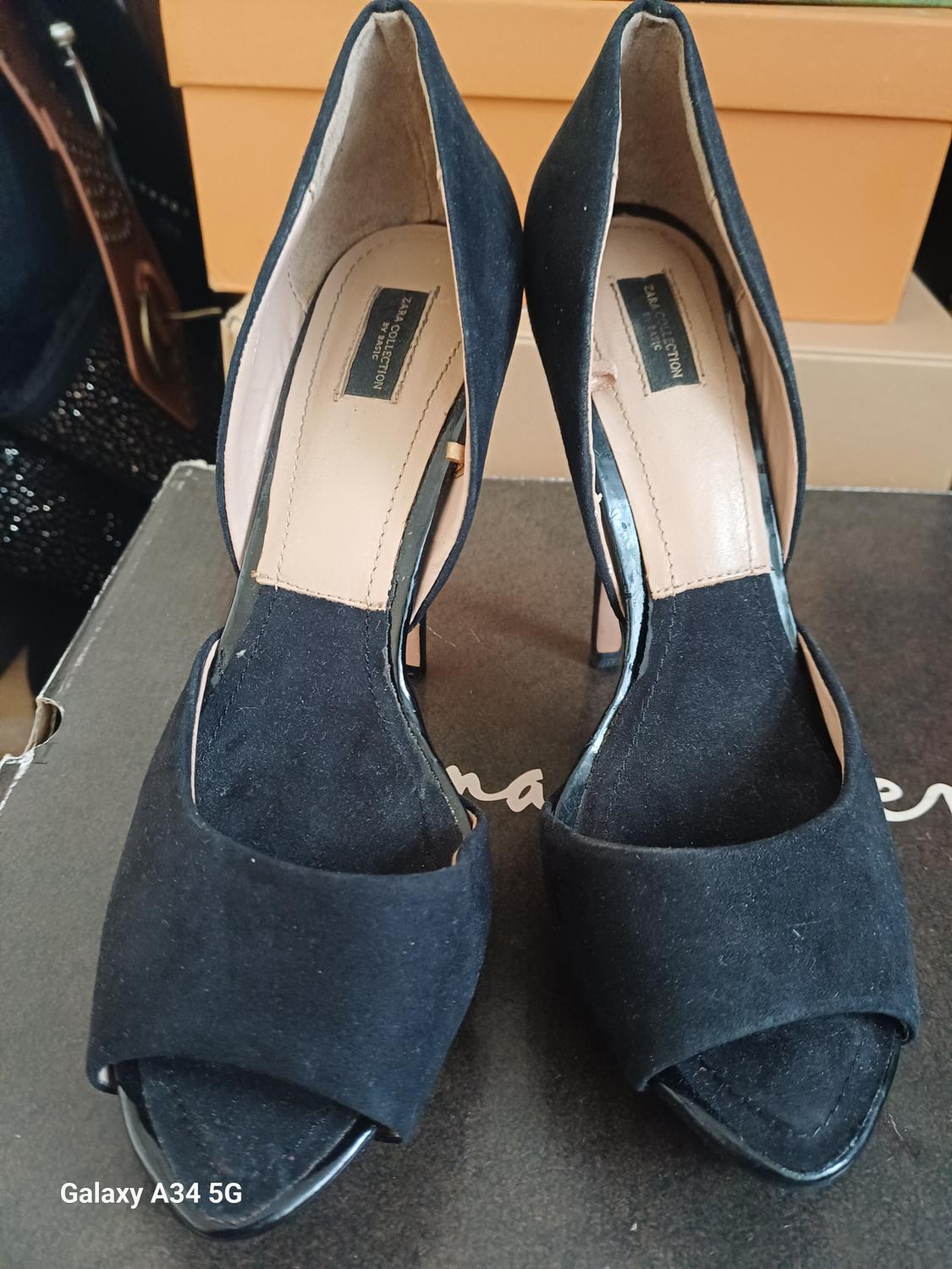 Front view of black high-heeled shoes with a visible label on the insole and a background of cardboard boxes.