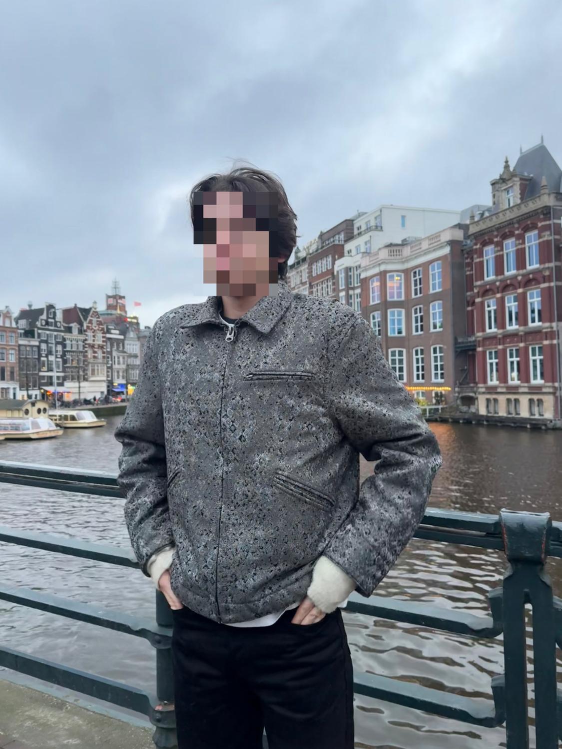 Model wearing a textured grey jacket and black trousers, posing on a bridge over a canal with boats and urban buildings in the background.