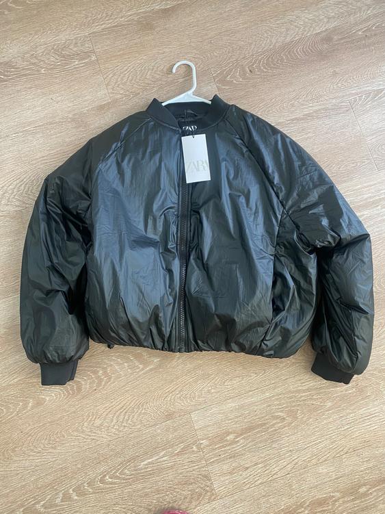 Black padded jacket with zipper and hang tag on white hanger on light wood floor.