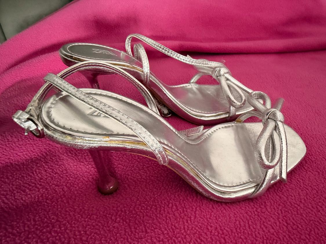 Silver high-heeled sandals with thin straps and a decorative bow, on a shiny pink fabric.