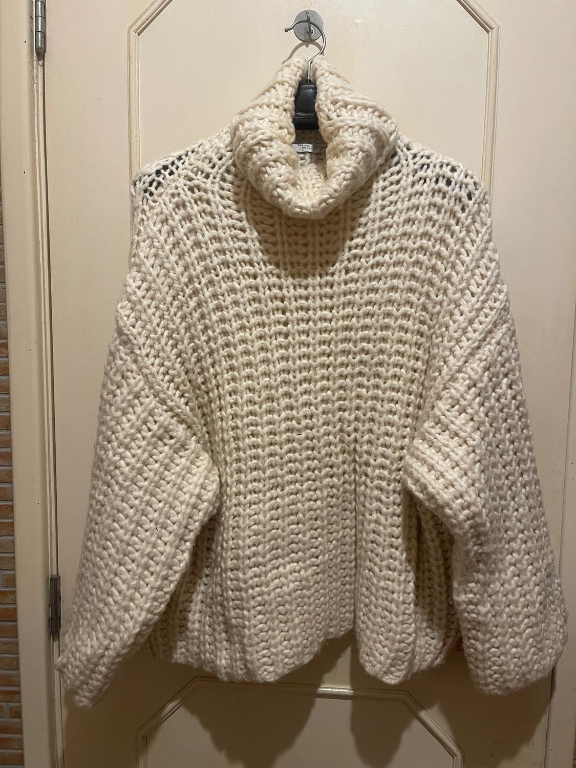 Chunky cream knitted jumper hanging on a door. High neck and long sleeve design, with visible knitted texture.