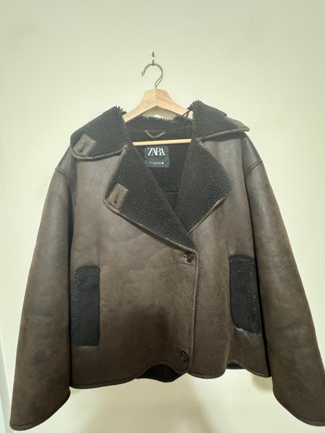 Brown jacket hanging on a wooden hanger against a light background. Wide lapels, textured dark lining inside and in the pockets. ZARA label.