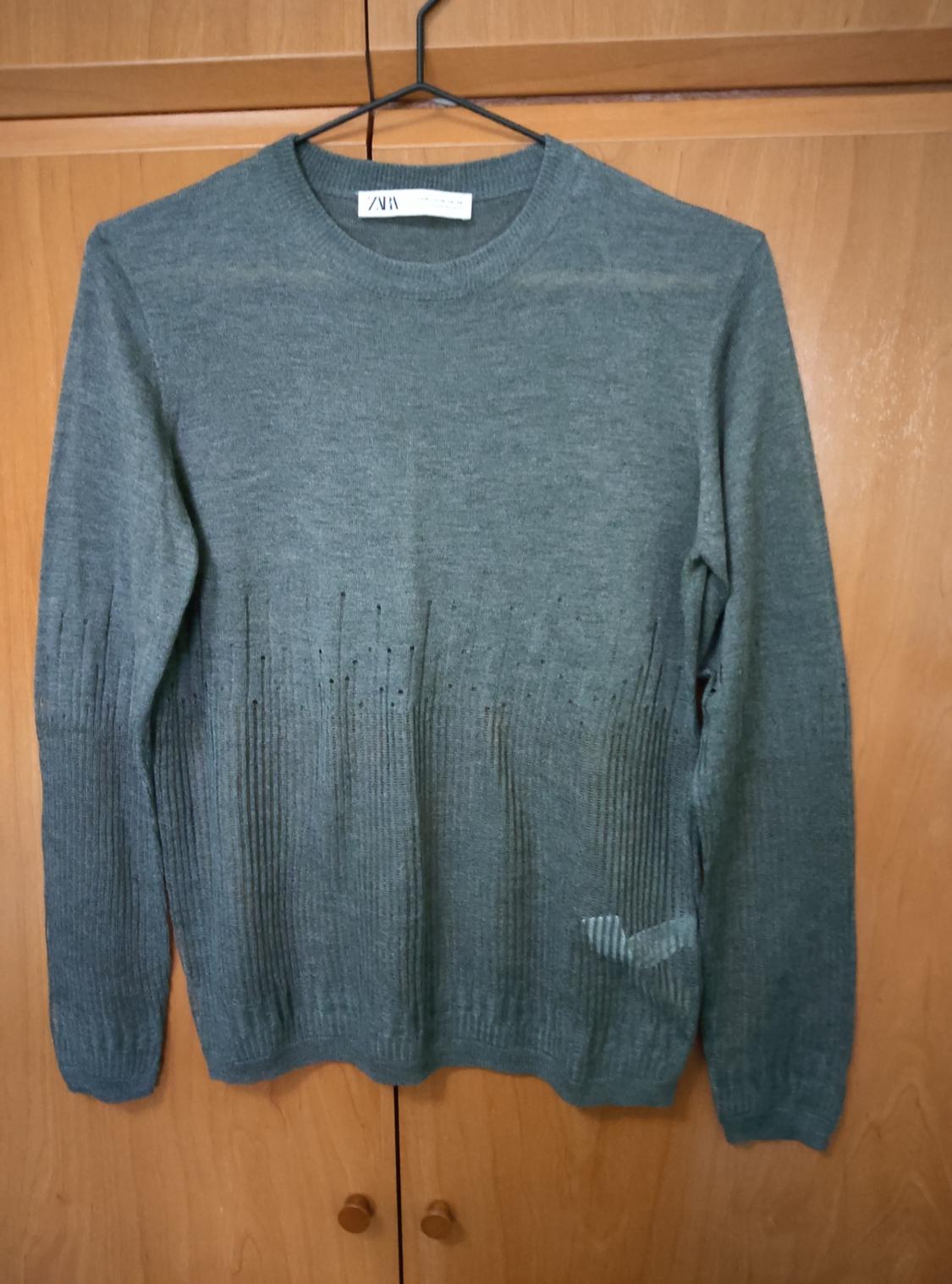Grey knitted jumper hanging on a hanger in front of a wooden wardrobe. Label visible on the collar.