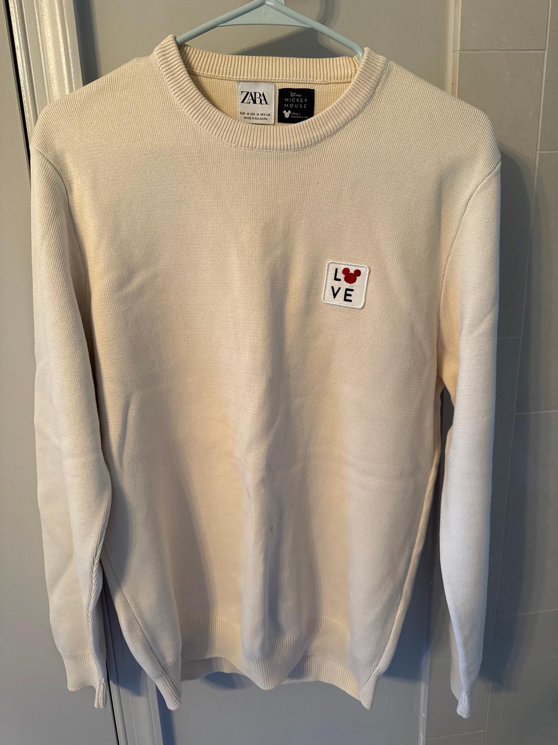 Beige jumper hanging on a hanger, with a 'LOVE' patch and a Mickey Mouse logo. Visible Zara and Disney tags.