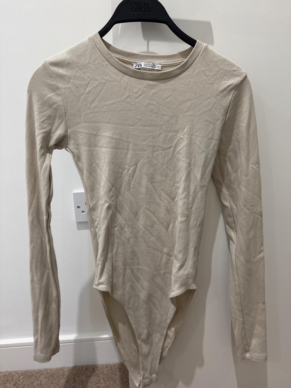 Beige long-sleeved bodysuit on a black hanger with a visible logo. White background and light brown carpeted floor.