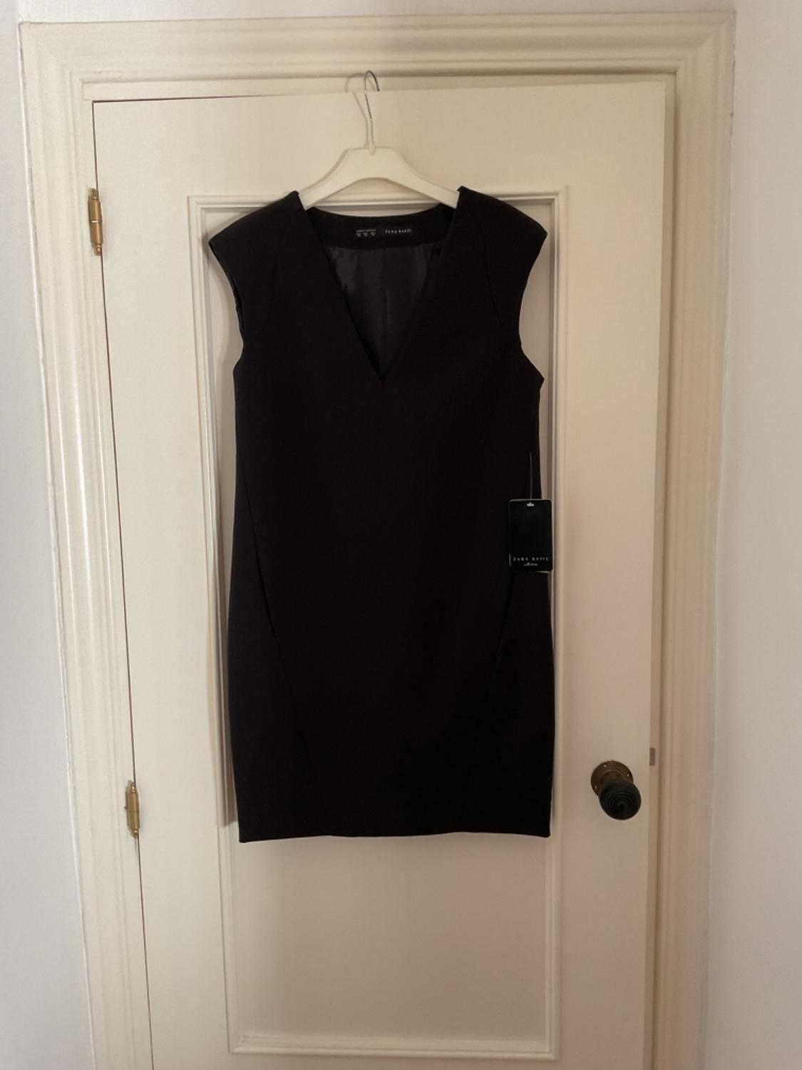 Front view of a sleeveless black dress with a V-neck and a hang tag.