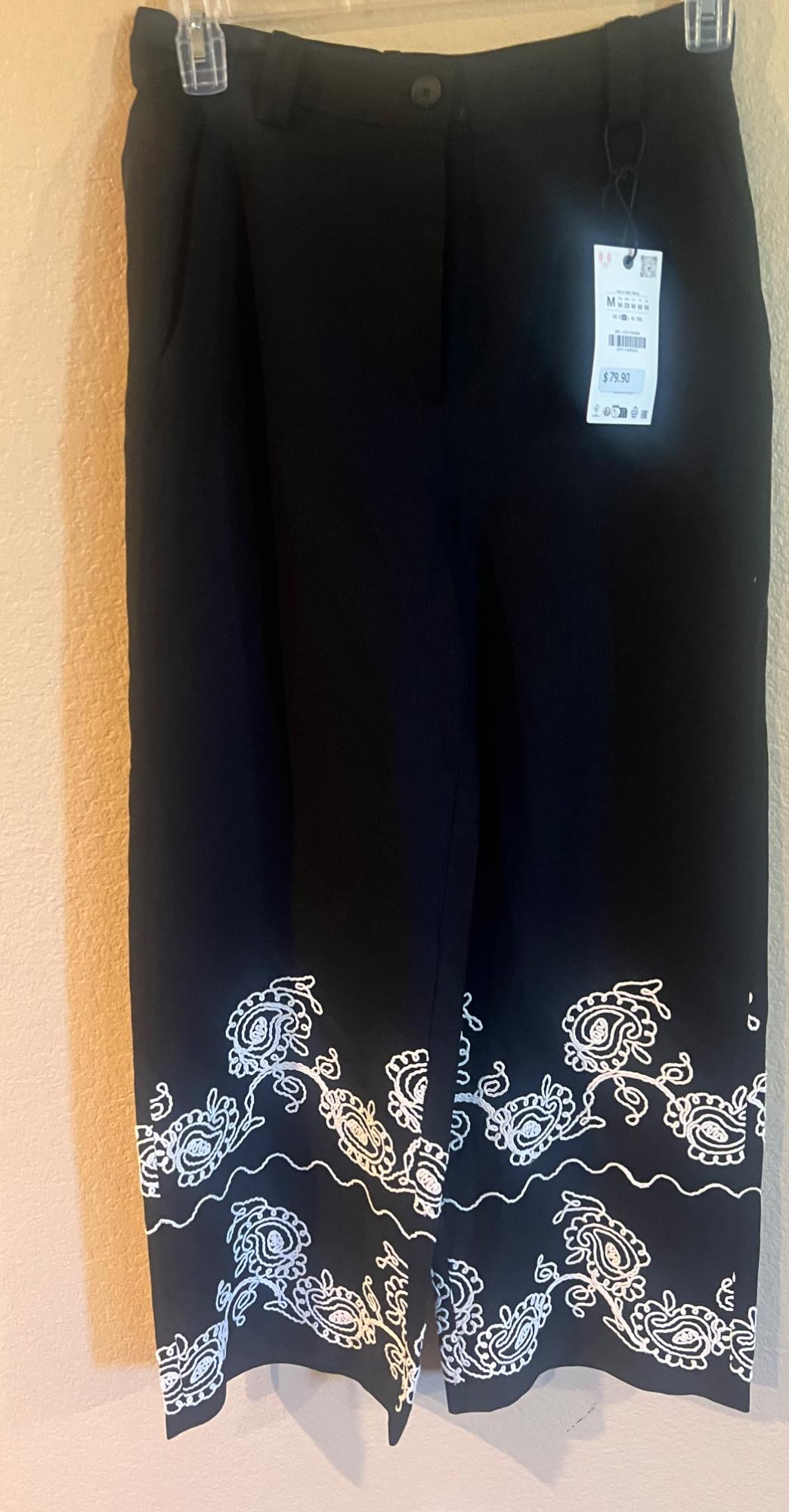 Black trousers with white embroidery on the hem, hanging on a coat hanger. Visible label with a price of $79.90.