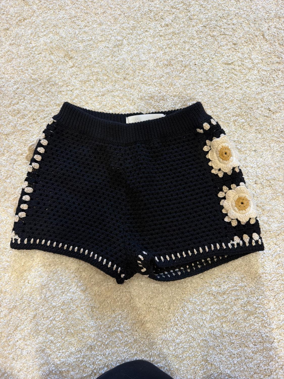 Black crochet shorts with white and yellow flowers on the side and contrast stitching, on a light-coloured carpet.