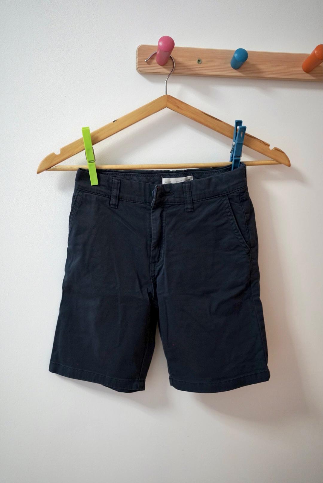 Navy shorts hanging on a wooden hanger, fastened with colourful pegs, against a white background.