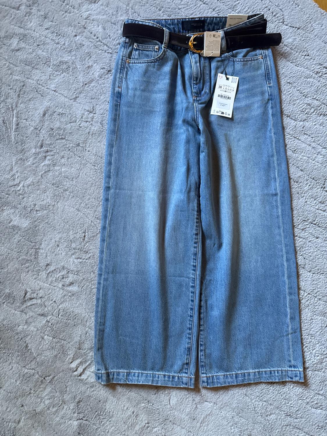 Light blue, wide-leg jeans with a black belt and gold buckle on a grey rug. Tags hanging from the belt.