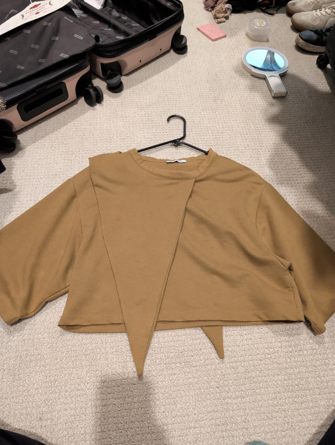 Brown long-sleeved T-shirt with an asymmetrical design on a beige rug, next to an open suitcase and a hand mirror.