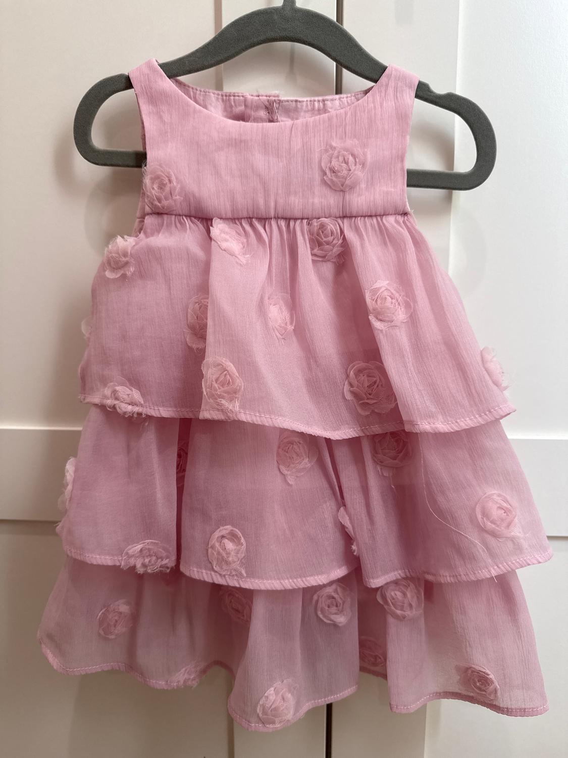 Girl's pink dress with ruffles and embossed flowers, hanging on a grey hanger against a white background.