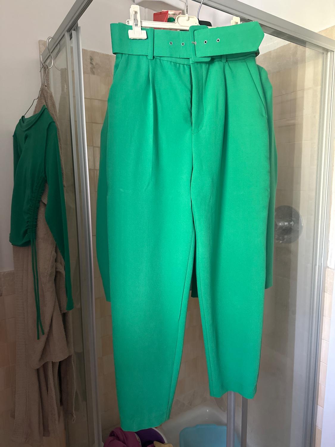 Green trousers hanging on a hanger inside a shower. In the background, a green garment and a beige towel on the wall.