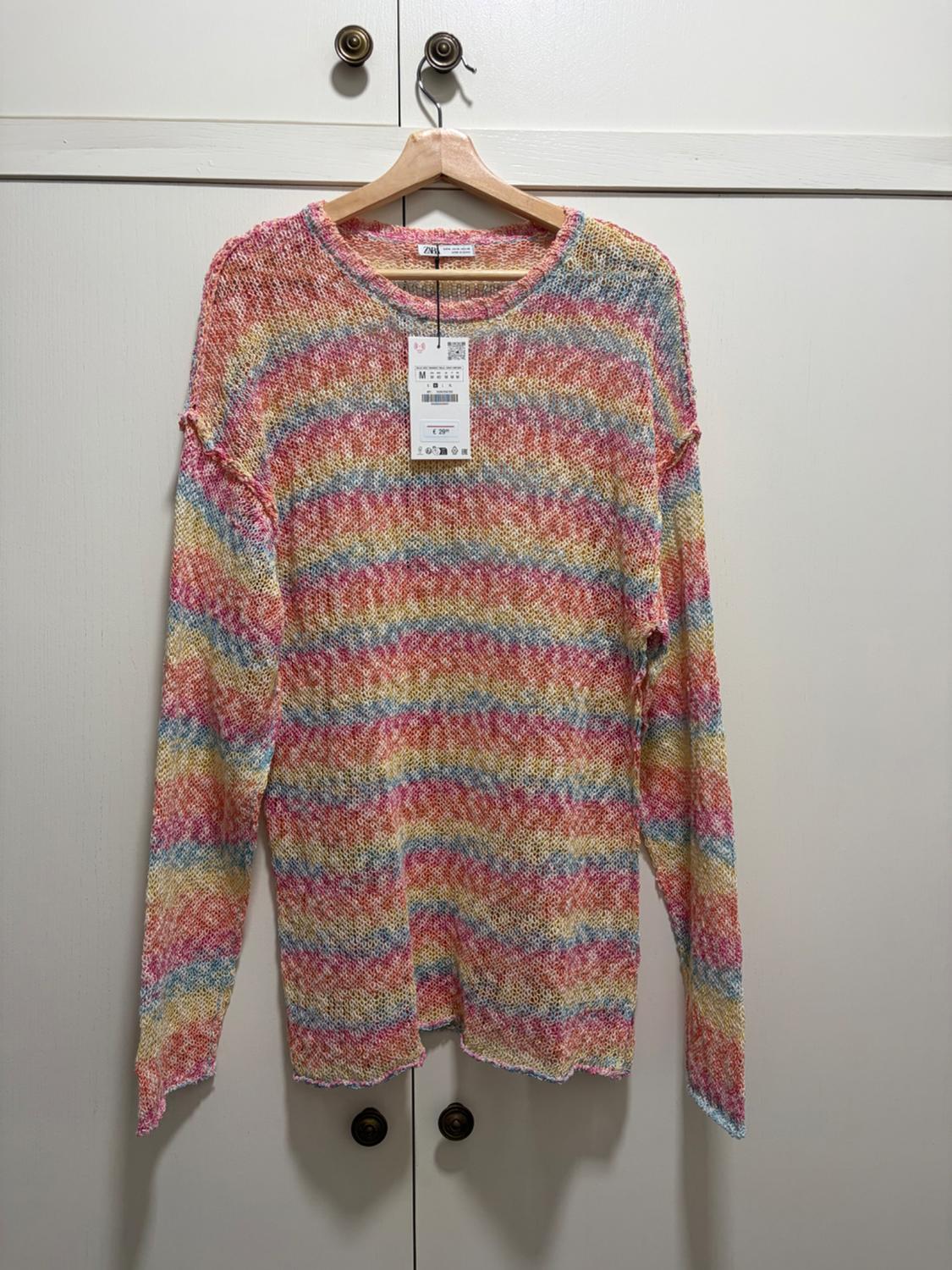Open-knit jumper with multicoloured stripes and a label, hanging on a wooden hanger in front of white wardrobe doors.