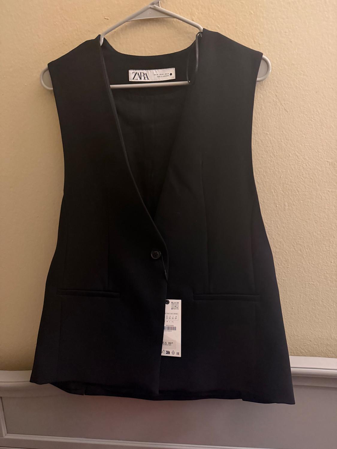 Black sleeveless vest hanging on a white hanger. Visible tag with the text "ZARA". Beige wall background.