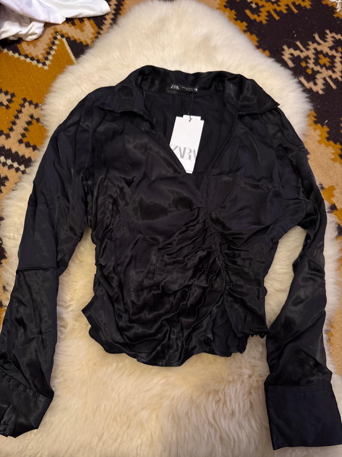 Black satin blouse with collar and long sleeves, visible label with text "ZARA", on a white rug and geometric background.
