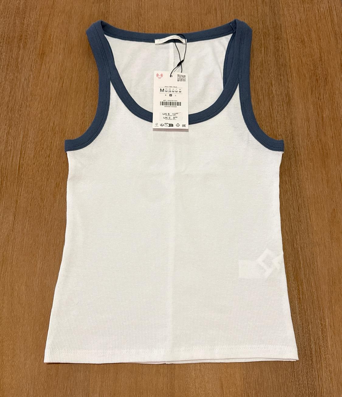 White ribbed tank top with blue trim on wood. Text: M US $ 12.90 UK £ 8.99.