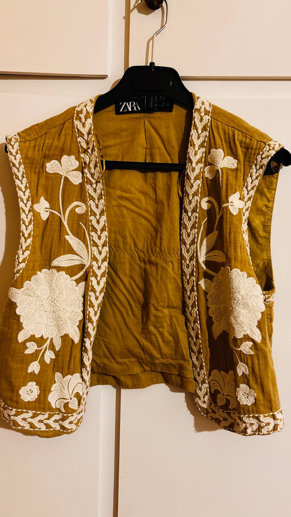 Mustard waistcoat with white floral embroidery, hanging on a black hanger with a "ZARA" label, against a white background.