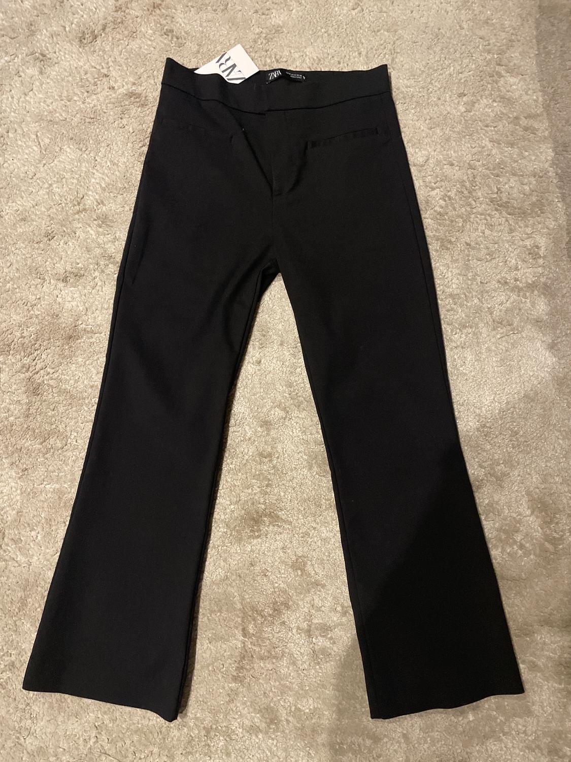 Black straight-leg trousers on a beige carpet, with a visible "Zara" label on the waistband.