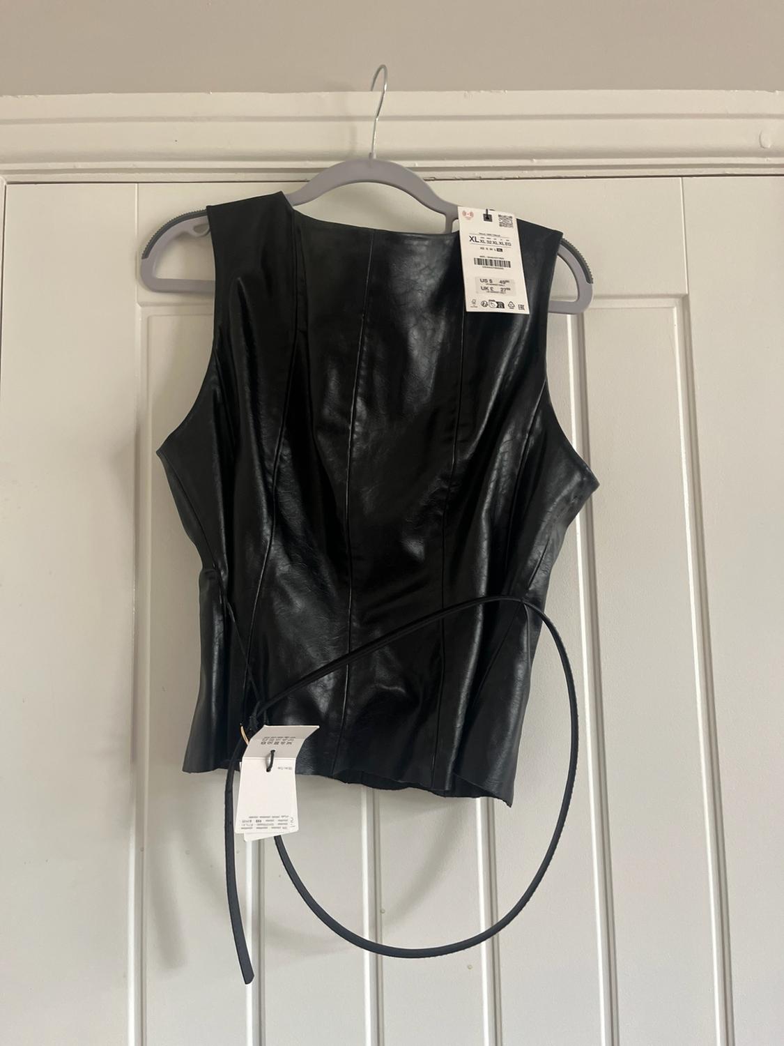 Black sleeveless top with a thin belt, hanging on a hanger over a white door. It has a visible label with the text XL.