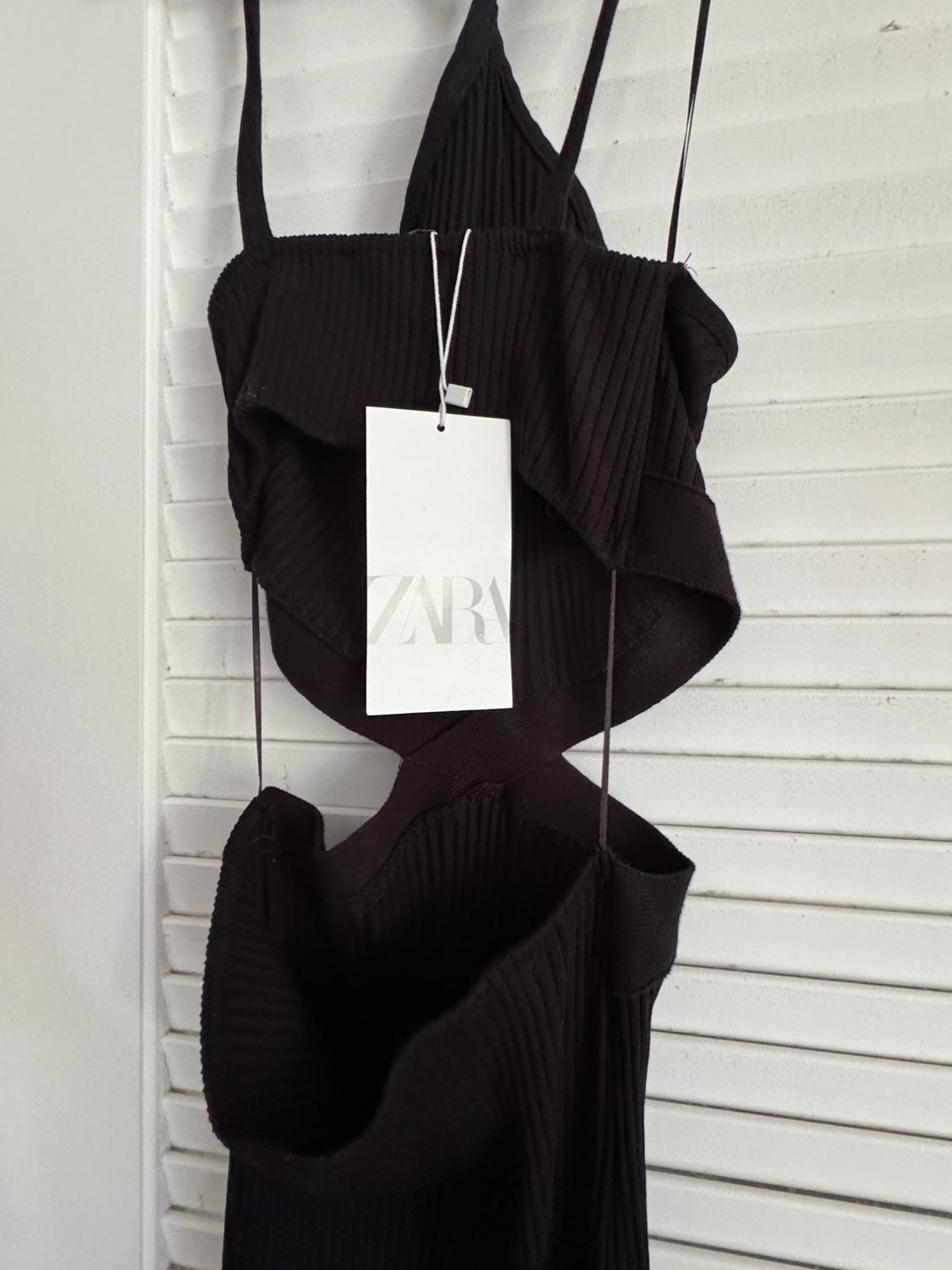 Sleeveless black dress with ribbed texture hanging on a white door. Visible label with the text "ZARA".