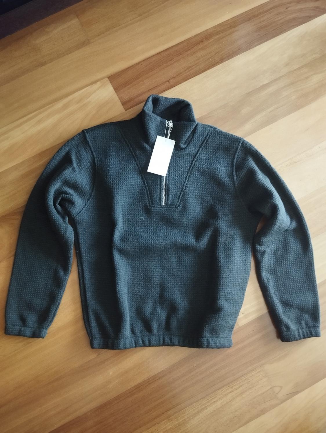 Dark grey jumper with a zip neck, placed on a light wooden floor. White tag hanging from the collar.