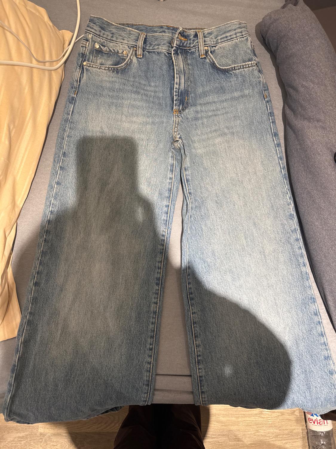 Straight-cut blue jeans on a grey bed, with pillows at the sides and an Evian water bottle on the floor.