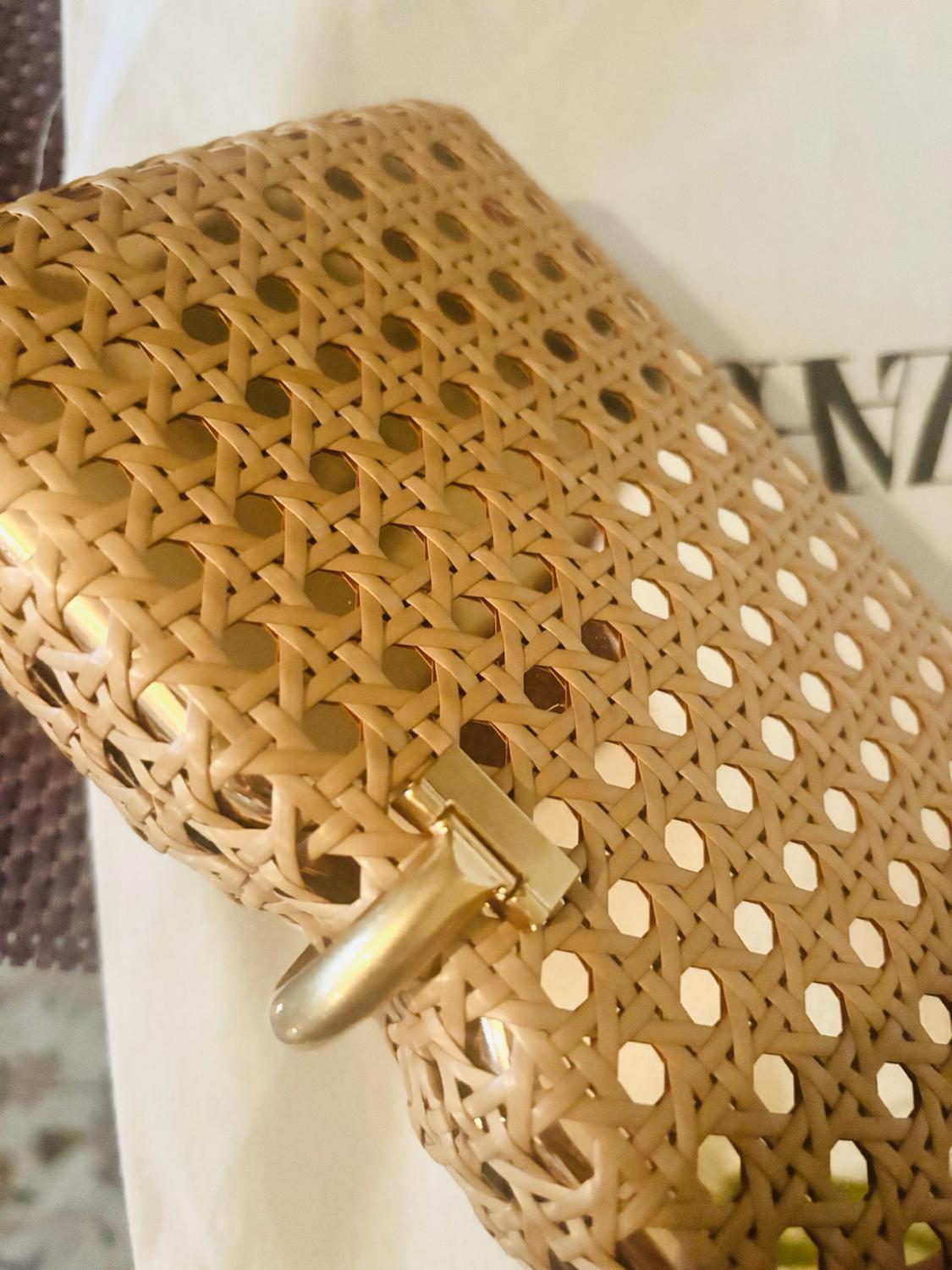 Detail of a beige netted braided-design bag with a gold metal fastening. In the background, a white surface with the partial text ZA.