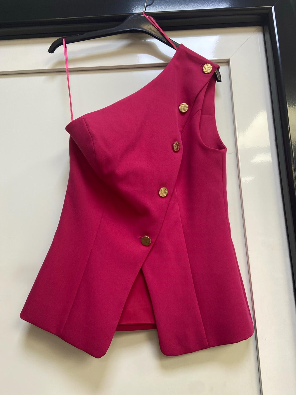 Asymmetrical fuchsia one-shoulder top with diagonal gold buttons, hanging on a black hanger against a white door.