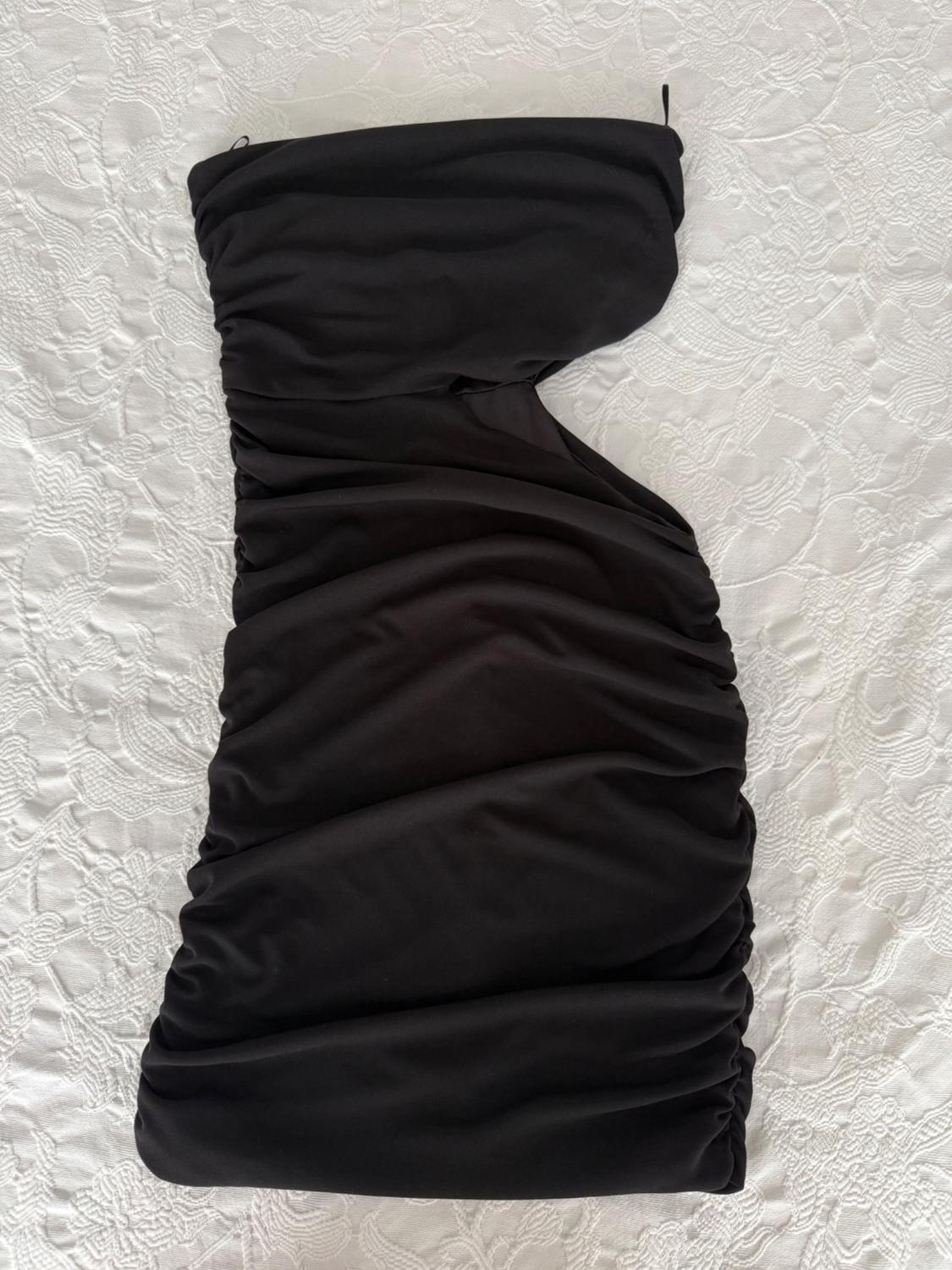 Ruched black mini dress, strapless with a side slit, laid out on a textured white fabric background.