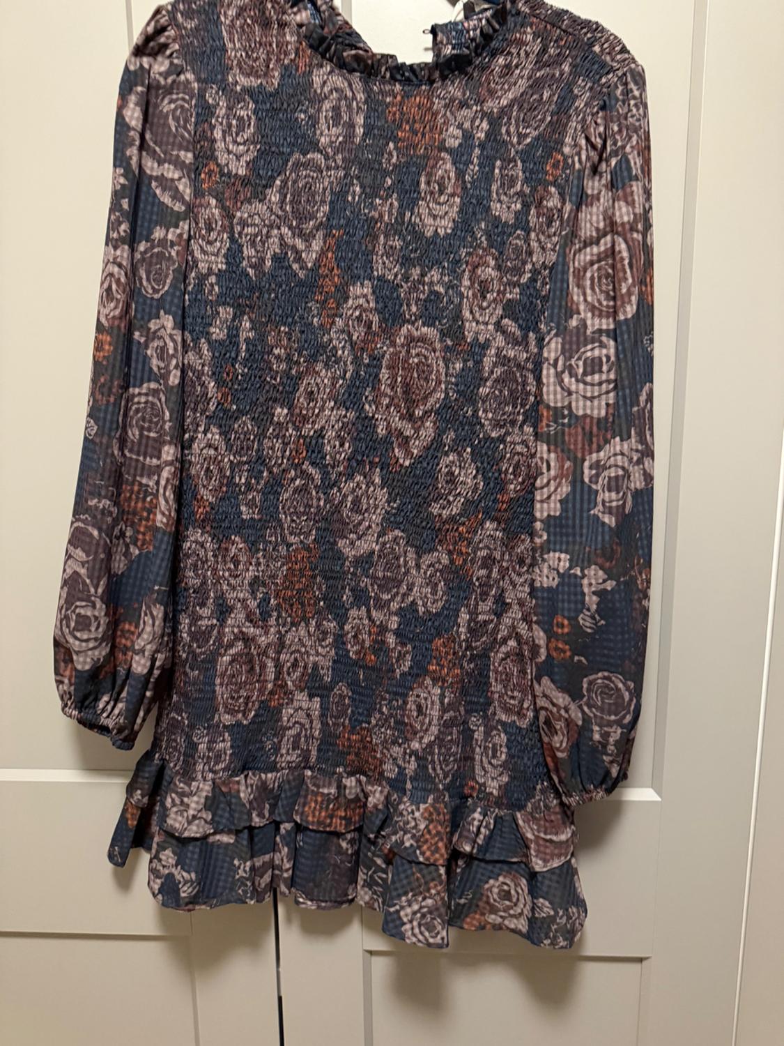 Short long-sleeve dress with a floral print, gathered bodice, and ruffle details at the neckline and hem, hanging on a white door.