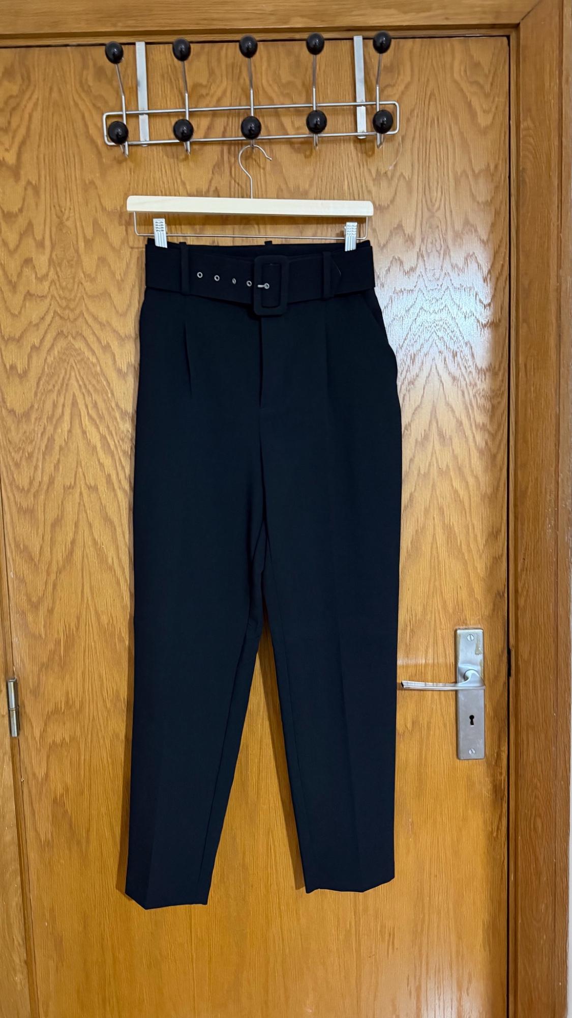Black tailored trousers with a matching belt, hanging on a hanger on a wooden door.