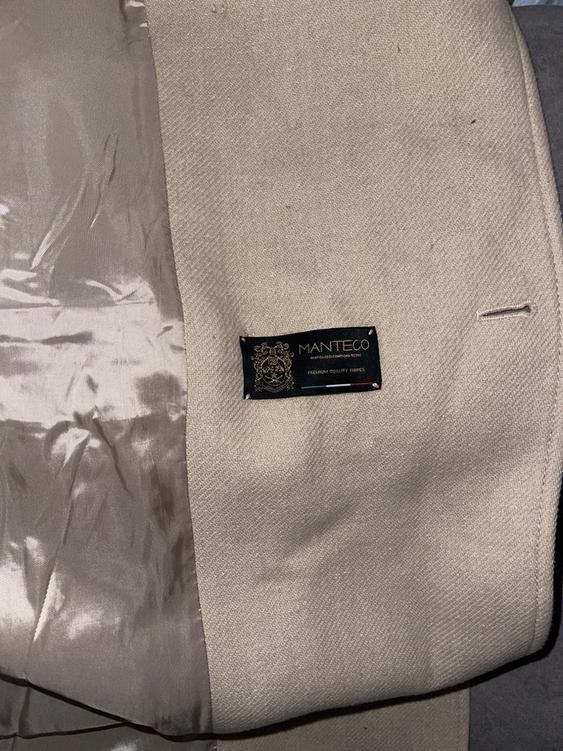 Black label "MANTECO" on beige fabric with satin lining. Text: "PREMIUM QUALITY FABRICS". Dark grey background.