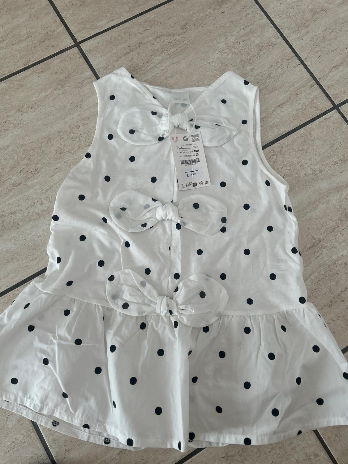 White, sleeveless blouse with black polka dots and bows on the front, on a tiled floor. Visible tag with product details.