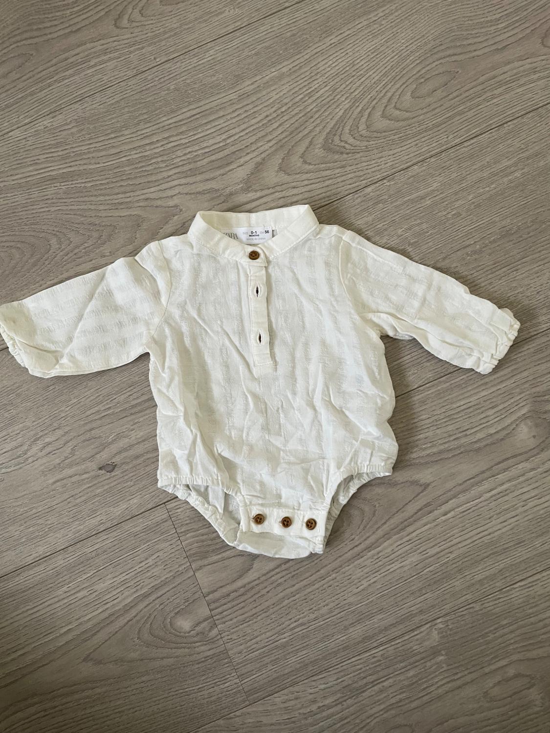 White baby bodysuit with long sleeves and brown buttons on the front and crotch, on a light wooden floor.