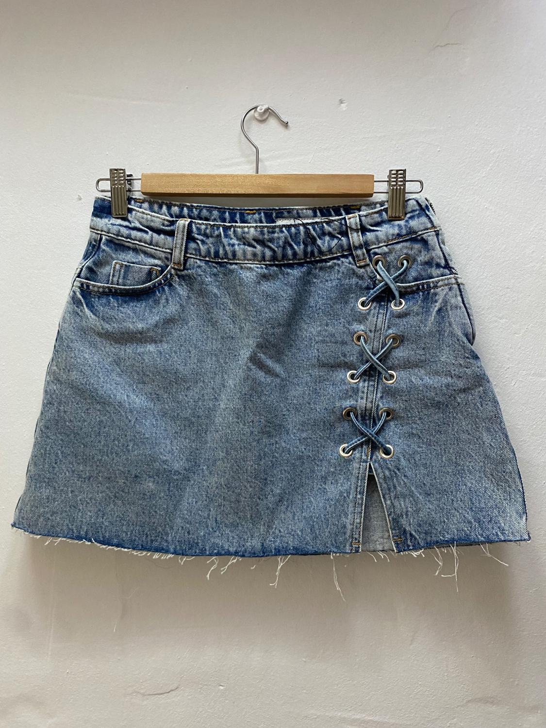 Blue denim mini skirt with crossover lace-up detail at the side, hanging on a wooden hanger against a white background.