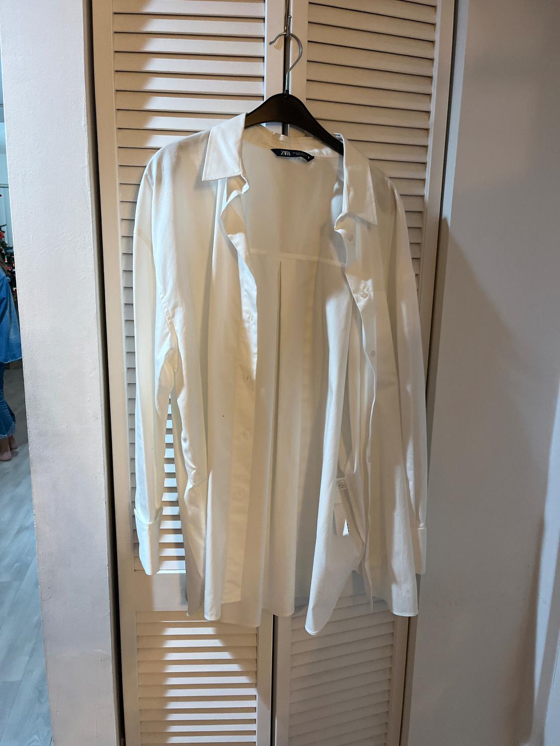 White long-sleeved shirt hanging on a black hanger in front of a white slatted door. Label visible on the collar.