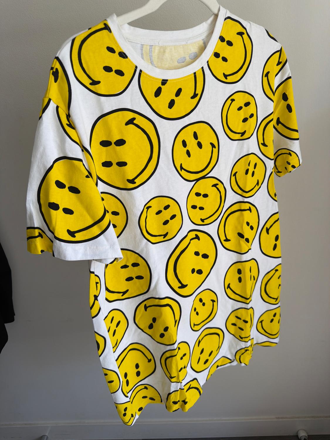 White T-shirt with yellow smiley face print, hanging on a hanger against a light grey background.