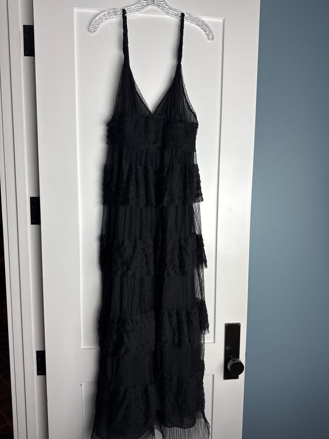 Long black tiered ruffle dress in semi-sheer fabric, hanging from a hanger on a white door.