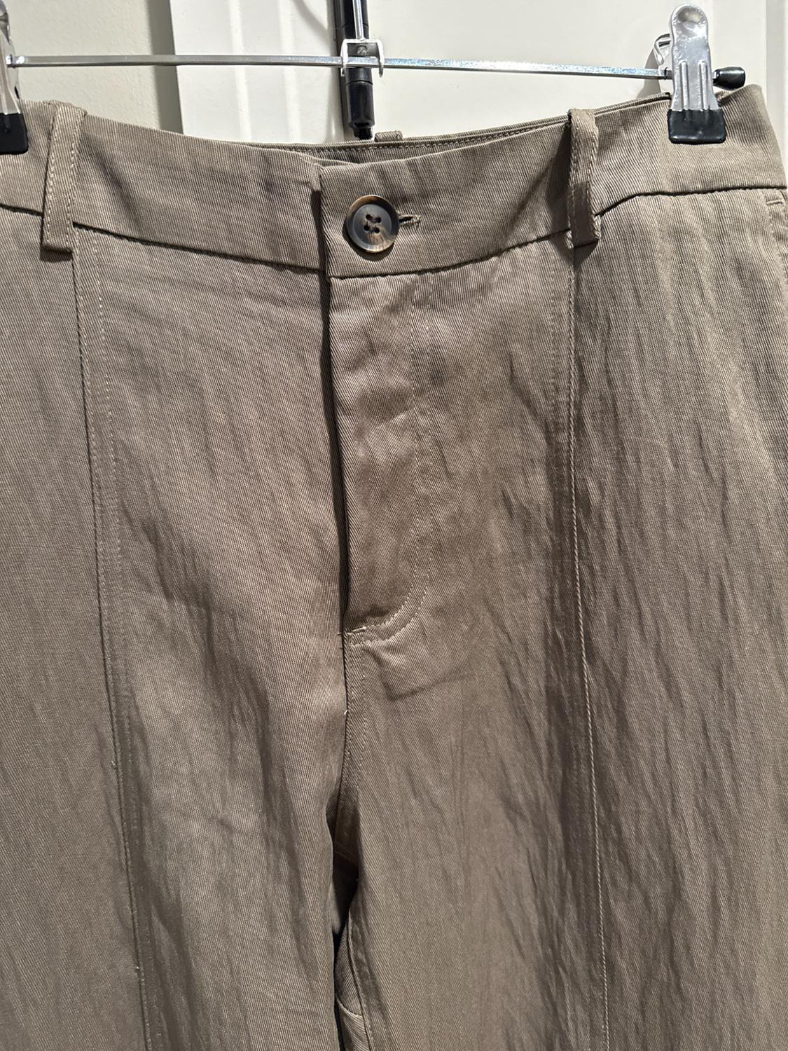 Front view detail of a pair of textured beige pants on a hanger. Shows the waistband with a dark button, belt loops, and vertical seams.