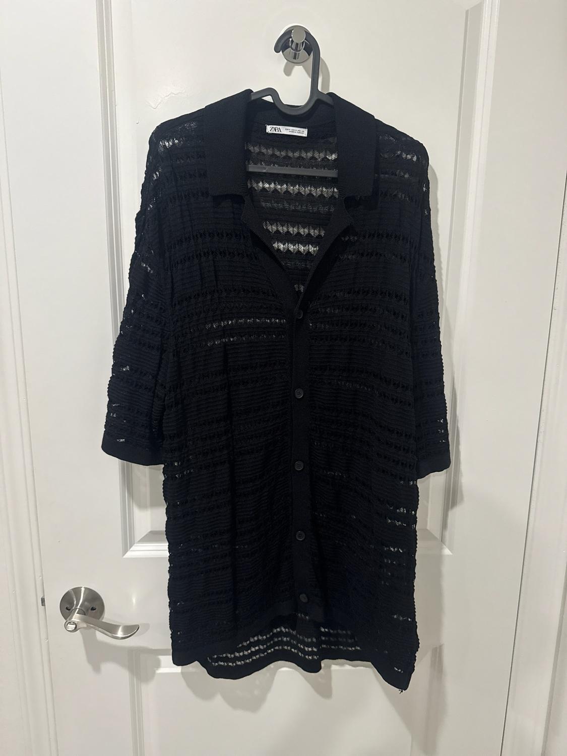 Black open-knit shirt with short sleeves and buttons, hanging on a hanger over a white door. ZARA label.