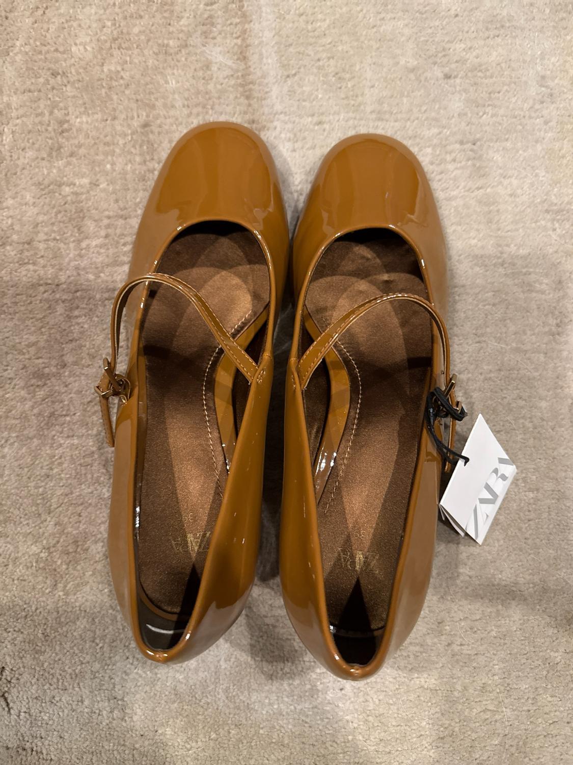 Brown patent leather Mary Jane shoes with a strap on a beige carpet. White insole and label with the text ZARA 39.