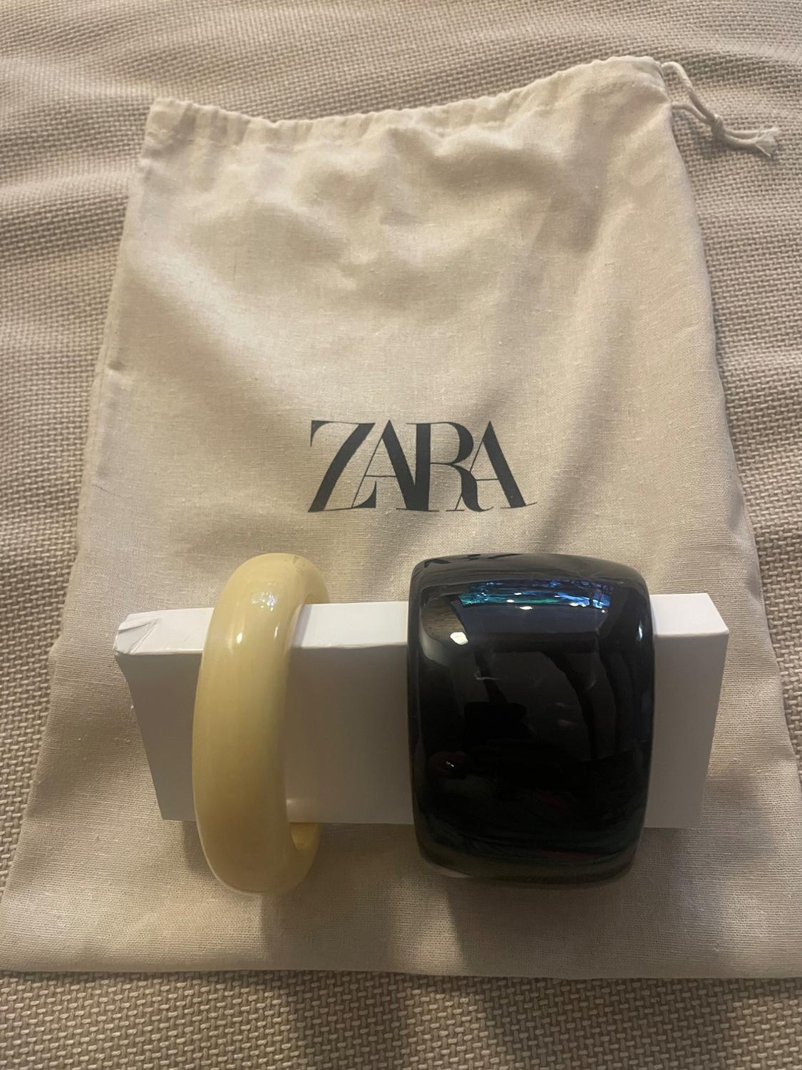 Two bracelets, one yellow and one black, on a fabric bag with the "ZARA" logo against a textured beige background.
